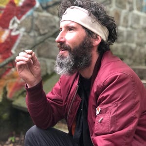 May include: A person with a beard and curly hair wearing a beige headband, a dark red bomber jacket with a zippered sleeve pocket, and a black shirt. The jacket features a small rectangular orange and white tag on the chest pocket area.