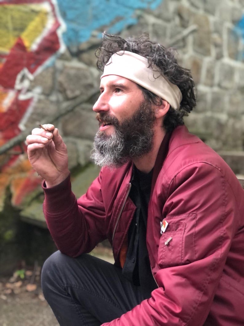 May include: A person with a beard and curly hair wearing a beige headband, a dark red bomber jacket with a zippered sleeve pocket, and a black shirt. The jacket features a small rectangular orange and white tag on the chest pocket area.