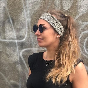 May include: A wide fabric headband featuring a black and beige leopard print pattern. The headband is worn across the forehead, securing long hair pulled back into a ponytail. The accessory is paired with round, dark-tinted sunglasses with thin metal frames.
