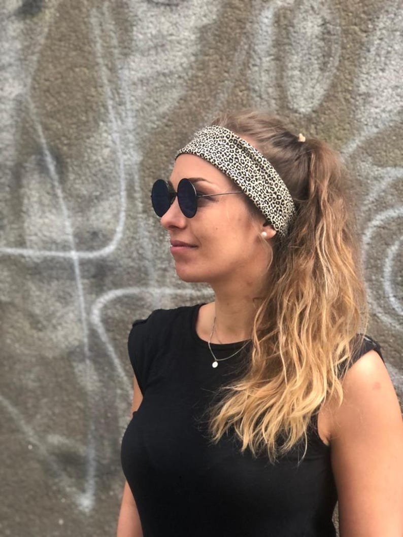 May include: A wide fabric headband featuring a black and beige leopard print pattern. The headband is worn across the forehead, securing long hair pulled back into a ponytail. The accessory is paired with round, dark-tinted sunglasses with thin metal frames.