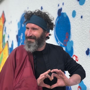 May include: A person with a beard wearing a dark teal headband, a black button-down shirt, and a maroon jacket draped over one shoulder. The person is forming a heart shape with their hands in front of their chest against a white wall with blue paint.
