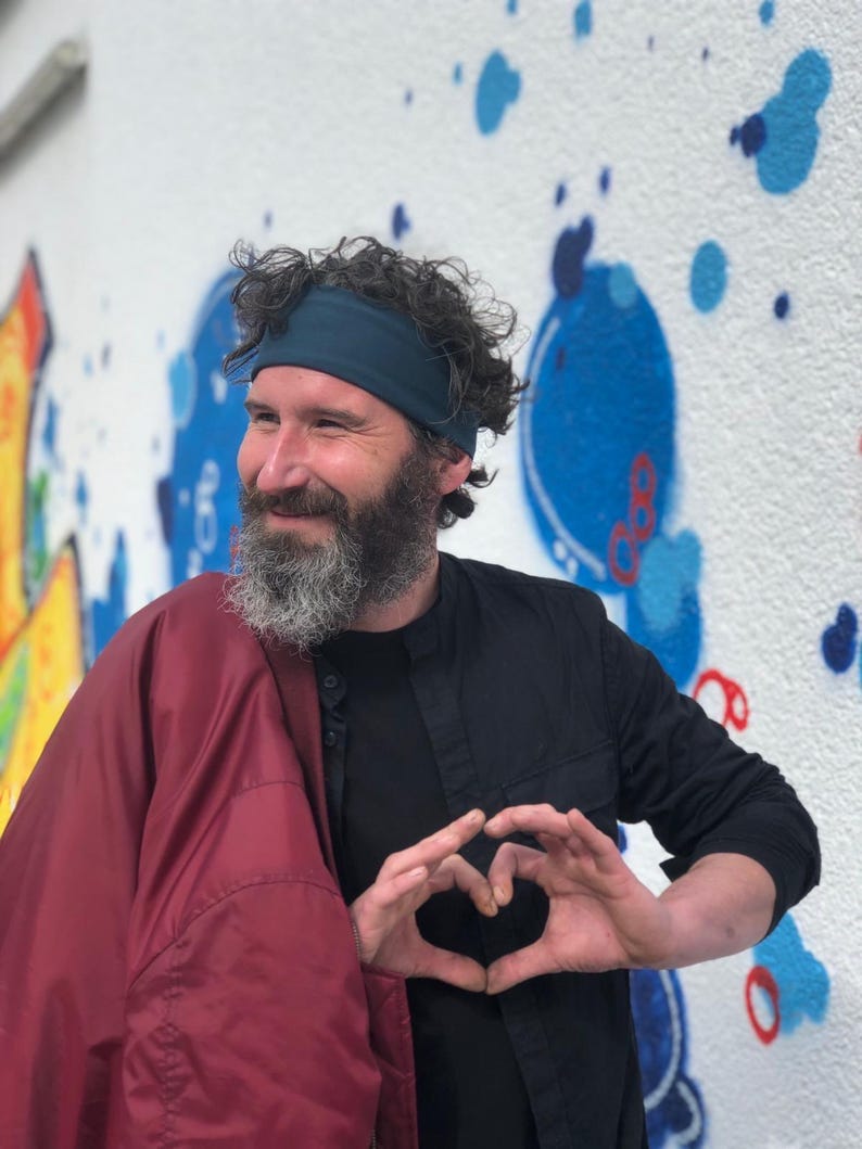 May include: A person with a beard wearing a dark teal headband, a black button-down shirt, and a maroon jacket draped over one shoulder. The person is forming a heart shape with their hands in front of their chest against a white wall with blue paint.