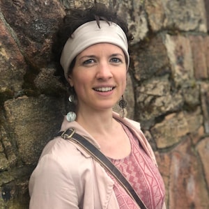 May include: A person wearing a light beige headband, a light pink jacket, and a pink patterned top. The person has dark curly hair and is wearing long, dangling earrings featuring circular, translucent, and dark metallic decorative elements.