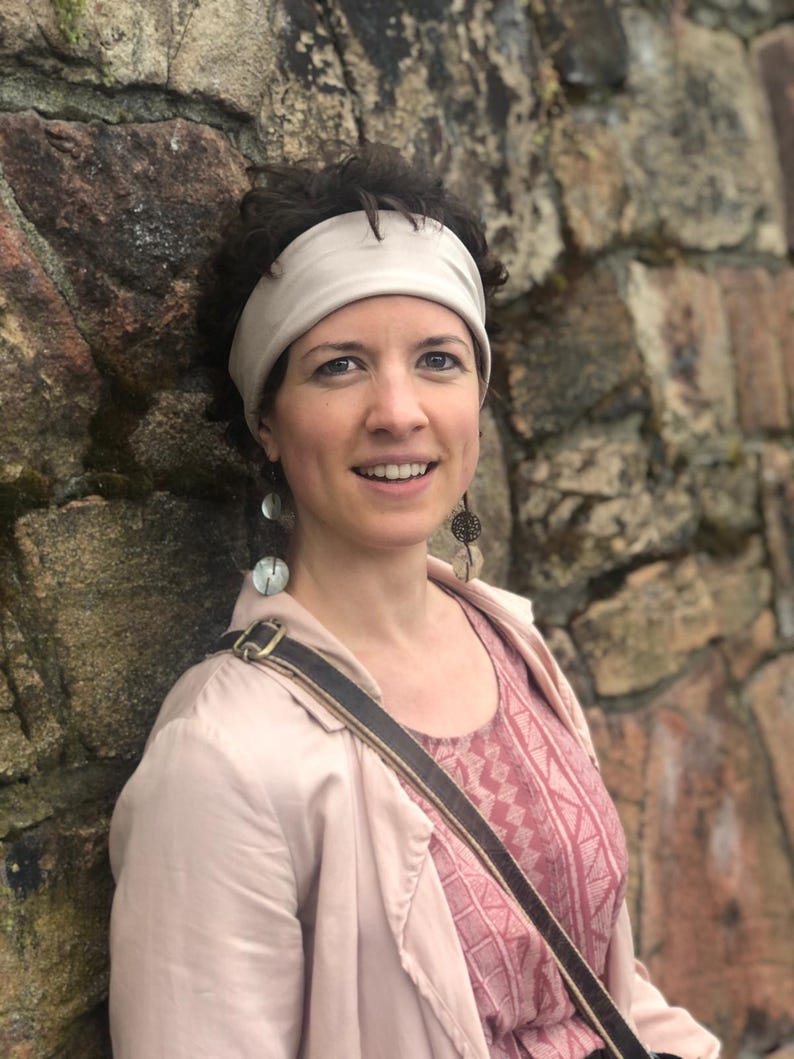 May include: A person wearing a light beige headband, a light pink jacket, and a pink patterned top. The person has dark curly hair and is wearing long, dangling earrings featuring circular, translucent, and dark metallic decorative elements.