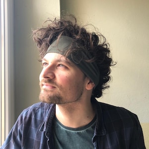May include: A solid olive green fabric headband worn across the forehead, featuring a wide, stretchy band design that sits against the hair. The headband is made of a smooth, matte material and provides a simple, functional accessory for hair.