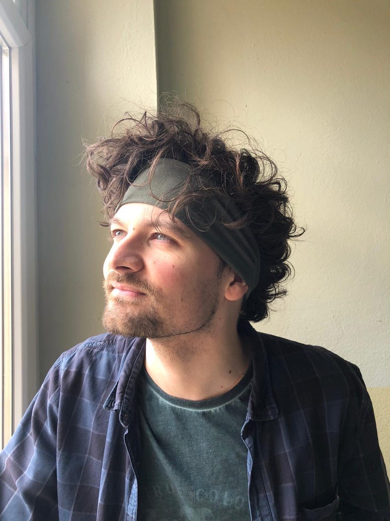May include: A solid olive green fabric headband worn across the forehead, featuring a wide, stretchy band design that sits against the hair. The headband is made of a smooth, matte material and provides a simple, functional accessory for hair.