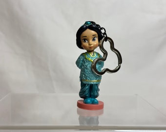 May include: A blue and gold figurine of a Disney princess with a teal head scarf and a silver key chain attached to her hand. The figurine is standing on a red base.