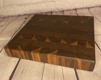 Könnte beinhalten: Ein dunkelbraunes Stirnholz-Schneidebrett. Das Brett ist quadratisch und besteht aus verschiedenen Holzstücken, wodurch ein Schachbrettmuster entsteht. Die Kanten sind abgerundet, und die Holzmaserung ist sichtbar.