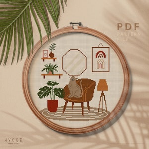 Puede incluir: Patrón de punto de cruz que representa una acogedora sala de estar con un gato sentado en un sillón, una lámpara, plantas y un espejo. El patrón está en un aro de madera redondo e incluye el texto "HYGGE CROSS STITCH" y "PDF PATTERN FILE".