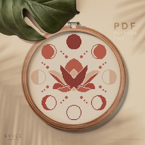 May include: A cross-stitch pattern in a wooden hoop features a floral design with moon phases in shades of red and peach. The text "PDF PATTERN FILE" is visible. The word "HYGGE" is printed at the bottom left.