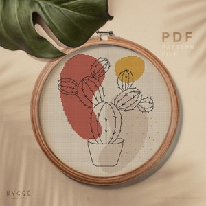 May include: A cross stitch pattern featuring a cactus plant in a pot with a red and yellow abstract background. The pattern is in a wooden embroidery hoop. The text "PDF PATTERN FILE" is in the upper right corner of the image. The text "HYGGE CROSS STITCH" is in the lower left corner of the image.