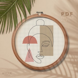 May include: A cross stitch pattern featuring a minimalist line drawing of a face with geometric shapes in shades of pink and brown. The pattern is displayed in a wooden embroidery hoop. The text "PDF PATTERN FILE" is displayed above the hoop. The text "HYGGE CROSS STITCH" is displayed below the hoop.