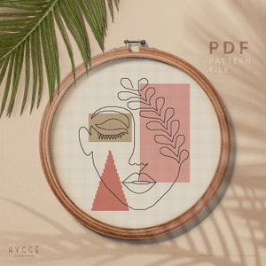 May include: A cross stitch pattern featuring a stylised face with a leaf design. The pattern is in a wooden embroidery hoop and the text "PDF PATTERN FILE" is visible at the top of the image.