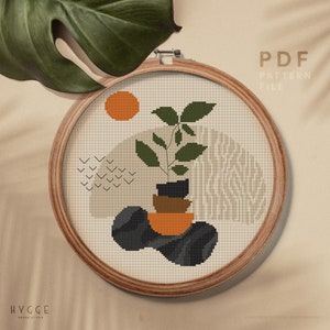 May include: A cross stitch pattern featuring a minimalist design with a plant in a pot, geometric shapes, and a sun. The pattern is in a brown wooden embroidery hoop. The text "PDF PATTERN FILE" is at the top of the image. The text "HYGGE CROSS STITCH" is at the bottom of the image.