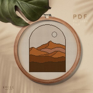 May include: A digital cross stitch pattern featuring a minimalist mountain landscape with a sun in the sky. The pattern is in a circular hoop and is labelled "PDF PATTERN FILE" and "HYGGE CROSS STITCH".