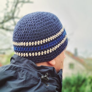 Op de afbeelding: Een marineblauwe gehaakte beanie met crème en blauwe strepen. De handgemaakte hoed is een close-up, die het getextureerde steekpatroon en de kleurdetails laat zien. De hoed is een stijlvol accessoire voor koud weer.