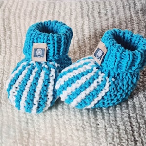 Op de afbeelding: Een paar blauwe en witte gestreepte gebreide babyschoentjes met een bruin label op een schoentje.