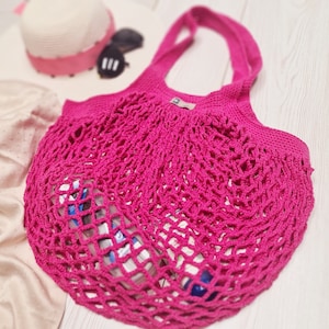 Könnte beinhalten: Eine leuchtend pinkfarbene gehäkelte Netztasche mit einem einzigen Trageriemen. Die Tasche ist aus einem feinen, zarten Garn gefertigt und hat eine lockere, offene Maschenstruktur. Die Tasche ist mit verschiedenen Gegenständen gefüllt, darunter eine Sonnenbrille.