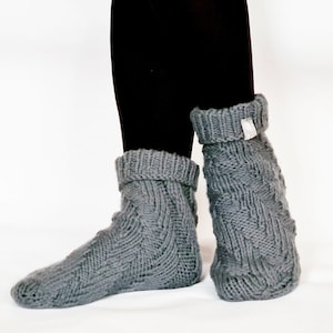 Puede incluir: Un par de calcetines de punto gris con un patrón de trenza. Los calcetines están doblados en la parte superior y tienen una etiqueta blanca en el puño.
