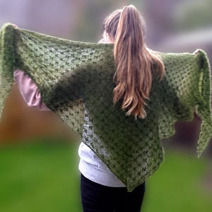 May include: A green knitted shawl with a delicate pattern is held open, showcasing its texture. The shawl is a muted green colour, with a slightly open weave. The background is a blurred green, suggesting an outdoor setting.
