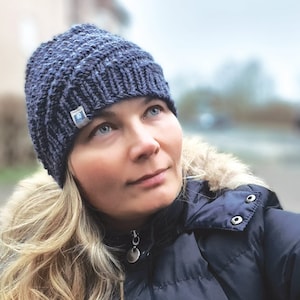 Könnte beinhalten: Eine Frau trägt eine blaue Strickmütze mit einem kleinen blauen Etikett an der Seite. Sie trägt eine dunkelblaue Jacke mit einem Pelzbesatz an der Kapuze. Sie schaut nach oben.