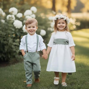 May include: Two children, a boy and a girl, are holding hands and smiling. The boy wears a white shirt, green pants with suspenders, and brown shoes. The girl wears a white dress with a green sash and a flower crown, and white shoes.