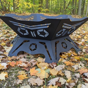 Puede incluir: Un brasero de metal negro con un diseño geométrico. El brasero está sentado sobre una cama de hojas caídas.