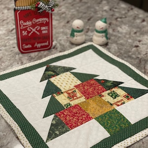 May include: A Christmas-themed quilted table runner with a tree design, featuring green, red, and gold fabrics. A red cookie jar with the text "Mrs. Claus Cookie Company" and two snowman salt and pepper shakers are also visible.