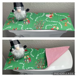 May include: A green Christmas-themed toilet seat cover with illustrations and text from a popular holiday story. The cover features a red and white striped underside and is paired with a gnome figurine.