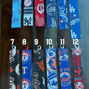 May include: Twelve fabric bookmarks with different baseball team logos. The bookmarks are numbered 1 through 12. The logos include the Cincinnati Reds, San Diego Padres, Los Angeles Dodgers, Texas Rangers, Detroit Tigers, New York Mets, and Boston Red Sox.