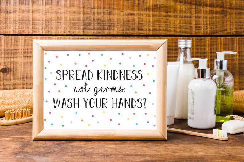 May include: A framed print with the text "SPREAD KINDNESS not germs. WASH YOUR HANDS!" is surrounded by colorful polka dots. The wooden frame sits on a wooden surface with soap, lotion, and a toothbrush.