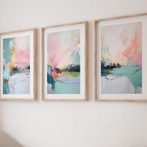 May include: A set of three vertical framed wall art prints featuring abstract compositions with blended washes of pastel pink, mint green, and soft blue, accented by thin black ink lines and gold leaf details, all housed in light wood frames.