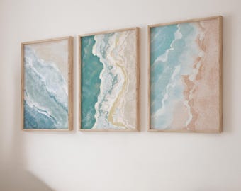 May include: A set of three rectangular wall art prints in light wood frames, each featuring an abstract aerial view of ocean waves meeting a sandy beach in shades of turquoise, blue, white, and beige, displayed side by side on a wall.