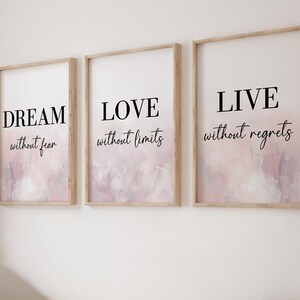 May include: Three vertical wall art prints in light wood frames. Each print features a white background with a soft pink and grey abstract watercolour wash at the bottom. The text reads DREAM without fear, LOVE without limits, and LIVE without regrets.
