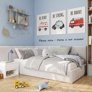 May include: Three rectangular wall art prints featuring watercolor illustrations of an ambulance, a police car, and a fire truck with the text BE KIND, BE STRONG, and BE BRAVE. The prints are displayed on a light blue wall above a bed in a room.