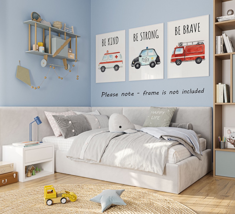 May include: Three rectangular wall art prints featuring watercolor illustrations of an ambulance, a police car, and a fire truck with the text BE KIND, BE STRONG, and BE BRAVE. The prints are displayed on a light blue wall above a bed in a room.