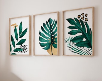 May include: A set of three framed wall art prints featuring botanical illustrations of dark green tropical leaves. The designs incorporate gold-colored accents, black zebra-like stripes, and a leopard print pattern on a clean white background.