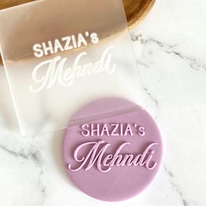 The image shows a personalized fondant embosser or stamp, for creating custom decorations on baked goods for a Mehndi ceremony. It features the custom text above the word "Mehndi" in a script font. These embossers are used to imprint designs and text onto fondant, icing, or dough, adding a personalized touch to cakes, cupcakes, and cookies for special occasions like weddings and Mehndi celebrations.