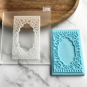 Puede incluir: Un molde rectangular transparente con un diseño rectangular blanco adornado y el texto "COSY BLOOM". Un jabón rectangular azul claro con el mismo diseño está al lado del molde. El diseño presenta una parte superior arqueada y un intrincado trabajo de volutas.