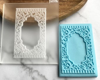 May include: A clear, rectangular mold with an ornate, white, rectangular design and the text "COSY BLOOM". A light blue, rectangular soap with the same design is next to the mold. The design features an arched top and intricate scrollwork.