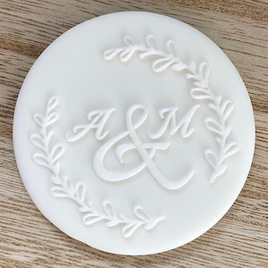 May include: White round cake topper with a white leafy wreath design and the initials "A & M" in a cursive font.