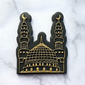 Può includere: Un tagliapasta nero e dorato a forma di moschea con due minareti e una cupola. Il tagliapasta è su una superficie di marmo bianco.