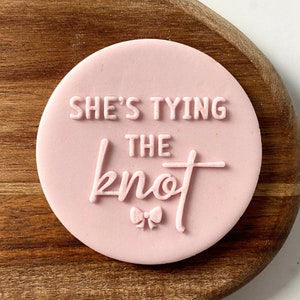 Può includere: Timbro rotondo per biscotti rosa con il testo "She's tying the knot" e un piccolo fiocco.