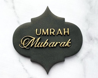 May include: A black and gold decorative cookie with the text "Umrah Mubarak" in gold lettering.