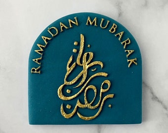 May include: A teal blue, rounded, decorative plaque with the text "Ramadan Mubarak" in gold lettering. The plaque features a gold Arabic calligraphy design in the center.