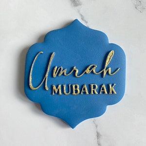 Può includere: Un oggetto blu a forma di ornamento con scritte dorate che recitano "Umrah MUBARAK". Il testo è in corsivo per "Umrah" e in stampatello per "MUBARAK". L'articolo è probabilmente un elemento decorativo o un biglietto di auguri.