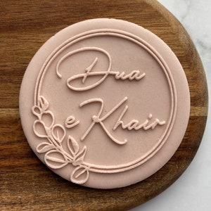 Puede incluir: Un sello de silicona rosa con el texto "Dua e Khair" en una fuente cursiva, rodeado de un círculo y un diseño floral.