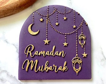 May include: Purple fondant cake topper with gold lettering that reads "Ramadan Mubarak". The topper features a crescent moon, stars, and hanging lanterns.