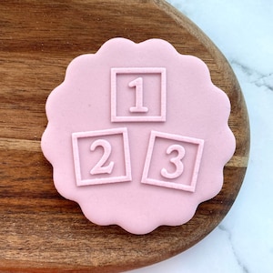 Può includere: Biscotto rosa in pasta di zucchero decorato con tre blocchi bianchi in pasta di zucchero con i numeri 1, 2 e 3.