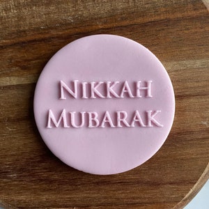 May include: A pink round cake topper with the text "Nikkah Mubarak" in white lettering.